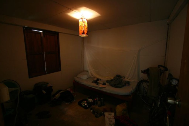 Ein Zimmer mit einem Bett in der Mitte, ein Fahrrad auf der rechten Seite, ein Fenster auf der linken Seite, ein Tischventilator und Taschen auf der linken Seite und Flaschen unten, mit Licht von oben.