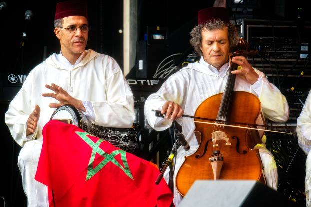 Zwei Männer in passenden Kostümen und Mützen, einer mit Brille, sitzen und spielen verschiedene Musikinstrumente, mit sichtbaren Musikanlagen dahinter.