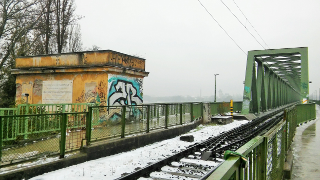 Eine schneebedeckte Zuggleis mit Graffiti an der benachbarten Wand, gesäumt von Geländern, Pfosten, Lichtern, Drähten und Bäumen unter einem sichtbaren Himmel.