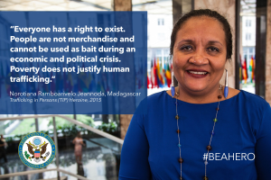 Eine Frau in einem blauen Kleid steht lächelnd vor einem Gebäude mit einer Aussage über Menschenhandel, mit Menschen, Pflanzen und Flaggen im Hintergrund.