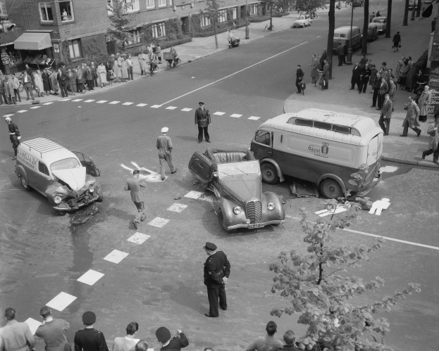 Schwarz-weiß-Szene eines Autounfalls an einer Straßenkreuzung mit mehreren Fahrzeugen, darunter ein Lieferwagen, und einer Gruppe von Schaulustigen; Gebäude, Bäume, Strommasten und Schilder mit Text sind im Hintergrund zu sehen.