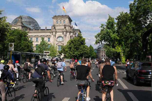 Eine Gruppe von Menschen, die auf Fahrrädern eine von Bäumen gesäumte Straße in Berlin, Deutschland, entlangfahren, mit Gebäuden auf der linken Seite und einer Bushaltestelle auf der rechten Seite unter einem bewölkten Himmel, eine Flagge sichtbar auf einem der Gebäude.