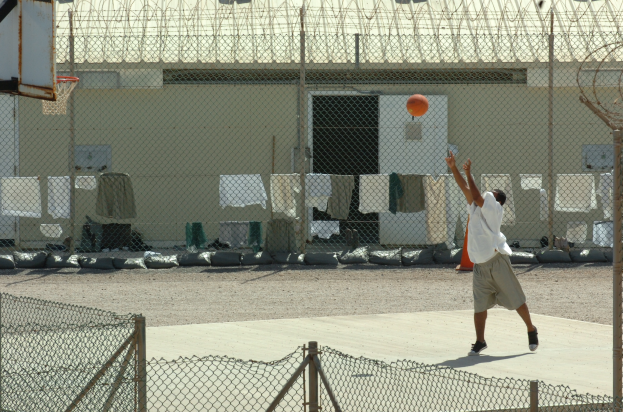 Ein Mann in Sportschuhen bereitet sich darauf vor, einen Basketballwurf vor einem Zaun zu machen, mit einem Basketballkorb links daneben und einem Gebäude im Hintergrund.