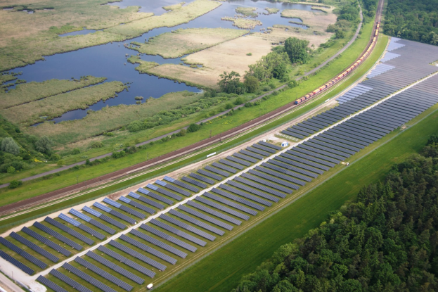 Luftaufnahme eines Solarparks mit Solarpanelen, umgeben von Bäumen, Gras, Pflanzen und Wasser, mit einem Zug auf einem nahen Bahngeleis.