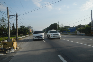 Zwei weiße Lieferwagen fahren auf einer Straße mit Strommasten, Kabeln, einer gelben Absperrung auf der linken Seite, Bäumen, Gebäuden und einem klaren blauen Himmel im Hintergrund.
