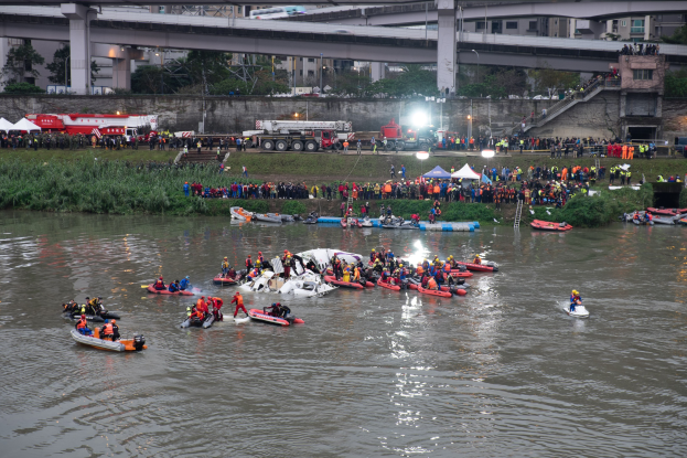 Eine Gruppe von Menschen in kleinen Booten auf einem Gewässer, die Rettungswesten tragen, während eines Rettungseinsatzes, umgeben von Vegetation, Zelten, Fahrzeugen, Infrastruktur und einer Brücke im Hintergrund.