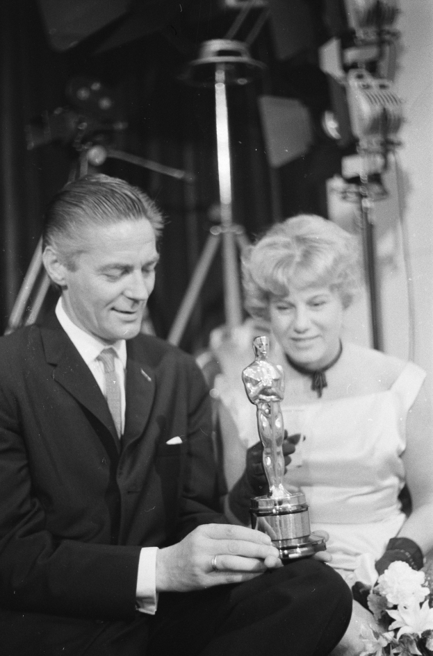 Ein Mann in einem schwarzen Blazer, weißem Hemd und Krawatte hölt eine Oscar-Statue, wa√hrend er neben einer Frau in Abendgarderobe sitzt, die einen Blumenstraußl in der Hand hölt, mit weicher Beleuchtung im Hintergrund.