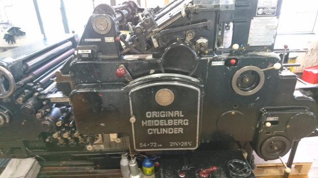 Eine Heidelberg Gto 52-4-2-2-Offsetdruckmaschine steht im Zentrum, umgeben von Flaschen und anderen Gegenständen auf dem Boden, mit Fenstern und einer Wand im Hintergrund.