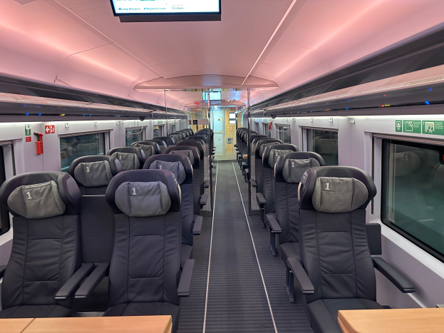 Das Innere eines Hochgeschwindigkeitszugs mit Sitzreihen und Tischen, Glasfenstern auf beiden Seiten und einem Bildschirm an der Decke.