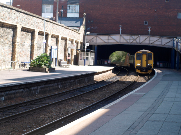 Ein Zug fährt auf Gleisen mit Plattformen auf beiden Seiten, eine Hauswand links, Straßenlaternen, Pflanzen, Bänke, eine Hütte, eine Brücke über die Plattformen und Gebäude und Masten im Hintergrund.