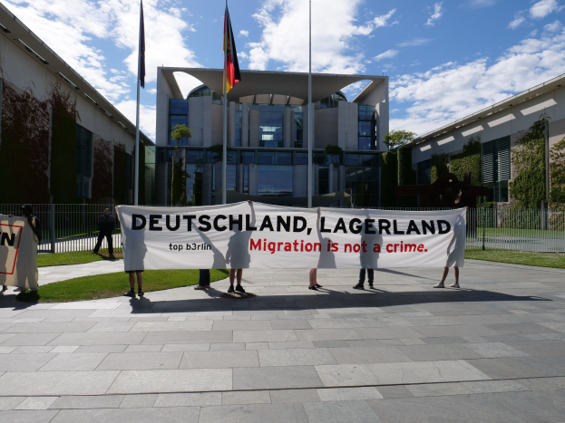 Eine Gruppe von Menschen mit einer Plakette, auf der 'Deutschland, Lagerland Migration ist kein Verbrechen' steht, vor einem Gebäude mit Fenstern, umgeben von Gras, einem Metallzaun, Pflanzen, Bäumen und Flaggen auf Ständen unter einem bewölktem Himmel.