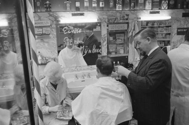 Schwarz-weißes Foto eines Mannes, der in einem Friseursalon einem Mann die Haare schneidet, mit einem Waschbecken, einem Spiegel, Regalen und einer Glastür im Hintergrund.
