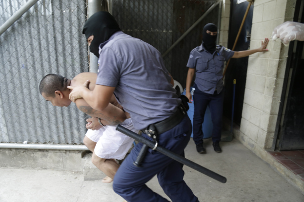 Ein Mann wird von zwei Polizeibeamten vor einem Gebäude eskortiert und von einem der Beamten festgehalten, während der andere einen Stock hält, mit einer Wand, einer Tür, einem Rohr und einem Netz im Hintergrund.