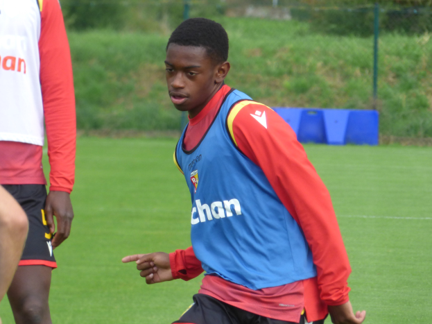 Ein junger Mann in einem blauen T-Shirt und schwarzen Shorts spielt Fußball auf einem grünen Feld, das von einem Zaun, Pflanzen und Bäumen umgeben ist.