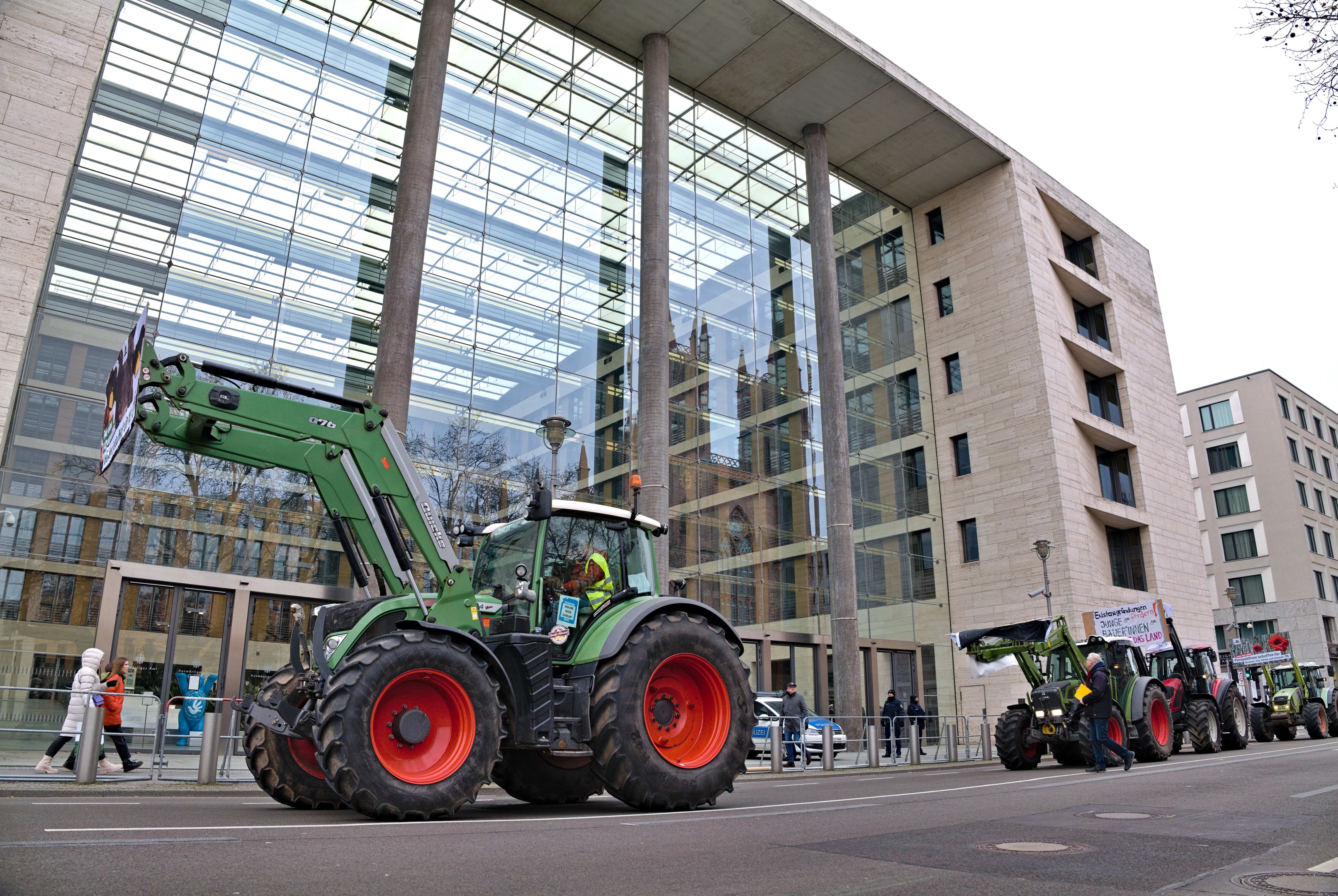 Eine Gruppe von Traktoren fährt eine Straße entlang vor einem Gebäude, mit Menschen auf dem Gehweg und einem Baum auf der rechten Seite, während es scheint, dass sie an einer Demonstration teilnehmen.