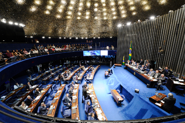 Eine Konferenzbühne mit sitzenden Personen an einem Tisch mit Papieren und Gegenständen, flankiert von Fahnen und einer Skulptur im Hintergrund, mit Sitzplätzen an Tischen und hinter einer Absperrung mit Bildschirmen, unter dekorativen Deckenleuchten.