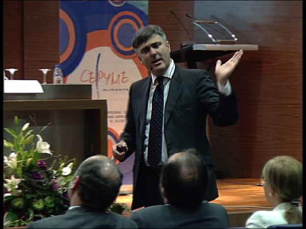 Ein Mann in einem schwarzen Blazer steht an einem Podium mit Mikrofonen, spricht in eines hinein, mit einem Blumentopf oder einem Strauß daneben und einem Tisch mit einem Buch, einer Wasserflasche und anderen Gegenständen in der Nähe; dahinter eine braune Wand und eine farbenfrohe Tafel mit Text, mit drei Personen, die im Vordergrund auf Stühlen sitzen.