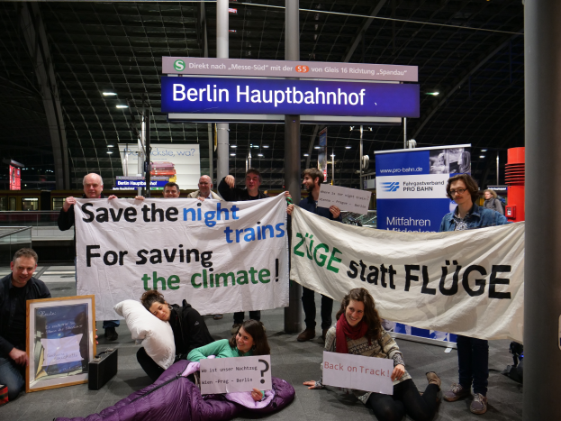 Eine Gruppe von Menschen sitzt auf dem Boden des Berliner Hauptbahnhofs und hält ein Banner mit der Aufschrift "Retten Sie die Nachtzüge, um das Klima zu retten". Es gibt Kissen, einen Fotoapparat und andere Gegenstände, die herumliegen. Im Hintergrund gibt es Schilder mit Text, Säulen, Lampen an der Decke und Geländer.