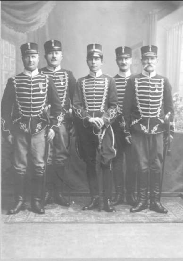 Eine Gruppe uniformierter Männer mit Mützen und Waffen steht zusammen, im Hintergrund eine Wand, ein Vorhang und ein Fenster; unten steht der Text "Sergei Ivanov und seine Offiziere, 1864".