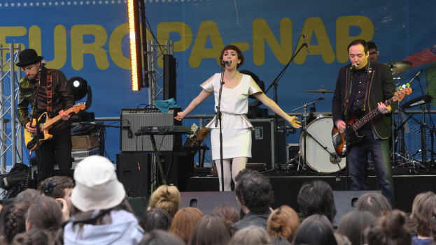 Ein Konzert mit vier Personen auf der Bühne: eine singende Frau in einem weißen Kleid in der Mitte, zwei Gitarristen links und rechts und eine Menge unten sowie Lautsprecher im Hintergrund.