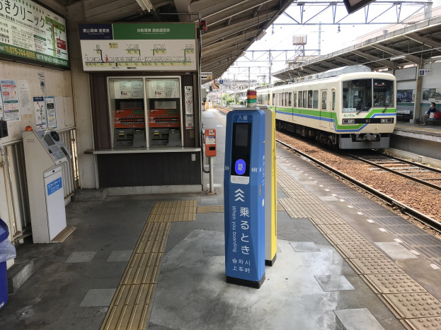 Ein Zug fährt in einen Bahnhof ein, mit Passagieren auf dem Bahnsteig und einem Automaten davor sowie einem Bildschirm und Metallstangen über dem Bahnsteig.