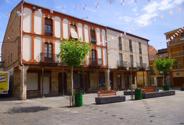 Ein Plaza Mayor mit Gebäuden mit Fenstern, Geländern, Bäumen, Bänken, Mülltonnen, Pfosten, Drähten und dekorativen Fahnen.