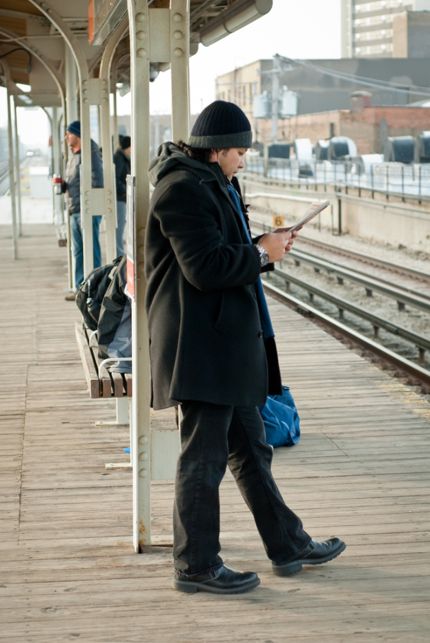 Ein Mann in einer schwarzen Jacke steht an einem Bahnhof mit einem Eisenstab und einer Hütte hinter ihm und Eisenbahnschienen und braune Gebäude rechts.