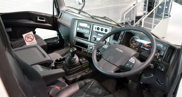 Ein Truck-Interieur mit einem Lenkrad, Armaturenbrett, Sitzen, Ganghebel, Tacho, Scheibenwischer, Seitenspiegel und weiteren Objekten, mit Geländern und weiteren Fahrzeugen im Hintergrund.