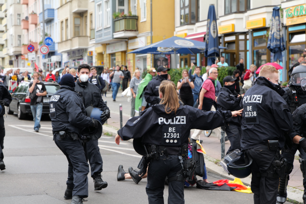 Eine Gruppe von Polizisten geht eine Straße entlang neben einer Menschenmenge, wobei einige Helme und Handschuhe tragen oder Schirme halten, mit Fahrzeugen, Schildern, Pflanzen und Gebäuden im Hintergrund.