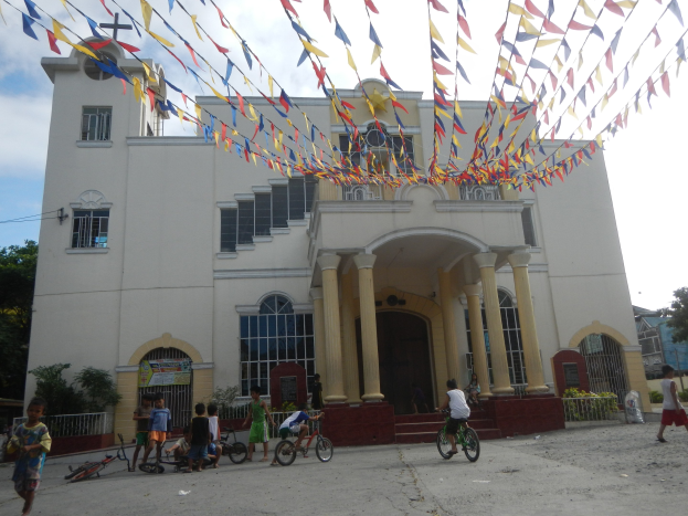 Eine Gruppe von Menschen, die auf Fahrrädern vor einer weißen Kirche mit architektonischen Details, Pflanzen, einem Zaun, einer Fahne, Bäumen und bunten Fahnen gegen einen bewölkten Himmel fahren.