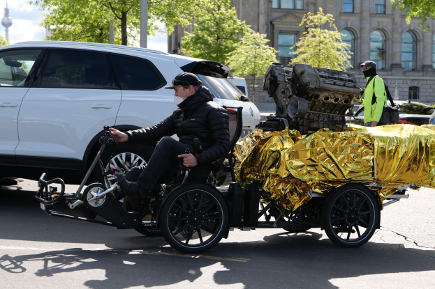 Ein Mann in einem Rollstuhl mit einem großen Motor an der Rückseite, umgeben von Fahrzeugen auf einer Straße mit Bäumen, Gebäuden und einem klaren blauen Himmel im Hintergrund.