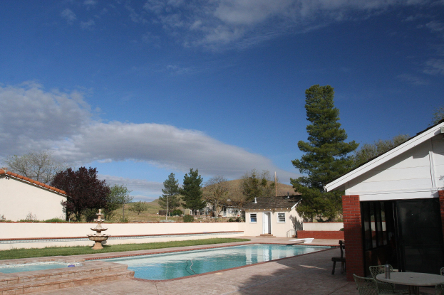 Ein Schwimmbad mit einem zentralen Brunnen, umgeben von grünem Gras und Bäumen, Gebäuden im Hintergrund, einem klaren blauen Himmel und einem Tisch mit Stühlen rechts daneben.