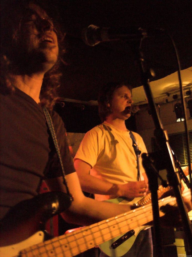 Eine Person in einem weißen Hemd singt in ein Mikrofon und spielt eine grüne Gitarre, während sie neben einer anderen Person in einem schwarzen Hemd und einer Brille steht, die ebenfalls singt und eine schwarze Gitarre spielt, mit Scheinwerfern und anderen Instrumenten im Hintergrund.