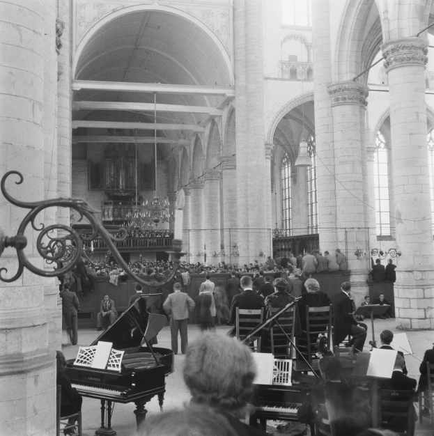 Ein Schwarz-Weiß-Foto eines Chores in einer Kirche, mit sitzenden und stehenden Menschen, einigen Musikern, einem Ständer auf der linken Seite und Säulen und einer Wand im Hintergrund.
